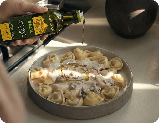 Hand drizzling Kofinas Basil Infused Olive Oil from the bottle over a bowl of pasta on a sunlit counter