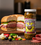 Jar of Classic Olive Muffalata beside a stacked deli sandwich with olives and peppers